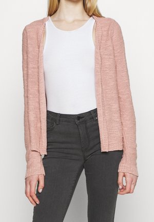 Cardigan lavorato a maglia rosa chiaro con apertura frontale, polsini a costine e vestibilità ampia, abbinato a una canotta bianca e jeans grigio scuro. La texture è morbida.