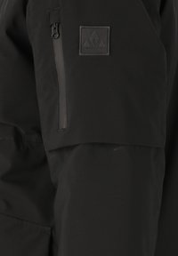 Mustat takki, jossa on vetoketjullinen tasku hihassa ja kankainen logo-patch. Materiaali on sileä ja matta.