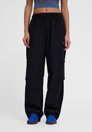 Person trägt lockere schwarze Hose, blaue Sneakers und ein kurzes graues Oberteil, steht vor einem weißen Hintergrund.