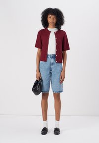 Cardigan corto bordeaux a maniche corte con un motivo texture, top bianco aderente, bermuda in denim azzurro chiaro e borsa nera. Le scarpe sono nere.