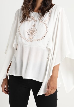 Woman wearing a loose white blouse with wide sleeves and beige floral embroidery on the chest, paired with black pants.