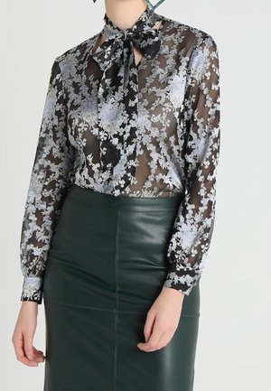 Ona ma na sobie przezroczystą czarną bluzkę z białymi kwiatowymi wzorami i wiązaniem pod szyją, zestawioną z ciemnozieloną skórzaną spódnicą z wysokim stanem.