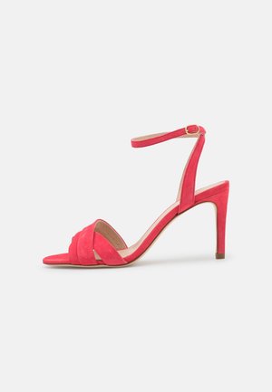 Sandale à talons hauts en daim rouge avec une sangle à la cheville et une boucle, bout ouvert et talon modérément fin sur fond blanc.