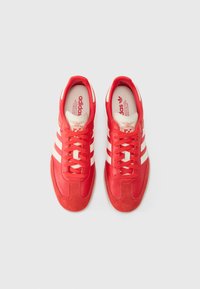 Røde sportsko med hvide striber, en overdel i tekstureret læder og ruskind, flade snørebånd og en gummisål. Har Adidas-branding.