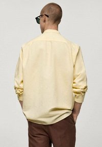 Gele, lange mouwen shirt gemaakt van een zachte stof, met opgerolde mouwen en een casual pasvorm, gecombineerd met bruine broek. Achteraanzicht getoond.