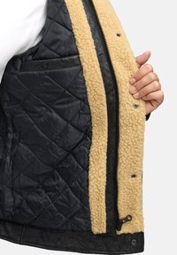Schwarze gesteppte Weste mit beigem Fleece-Futter, ausgestattet mit einer Reißverschlusstasche, Druckknöpfen und einem strukturierten Lederabschluss.