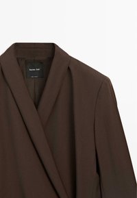 Donkerbruine blazer met sjaalkraag, zichtbare Massimo Dutti-label binnenin, gemaakt in Marokko, afgebeeld tegen een witte achtergrond.