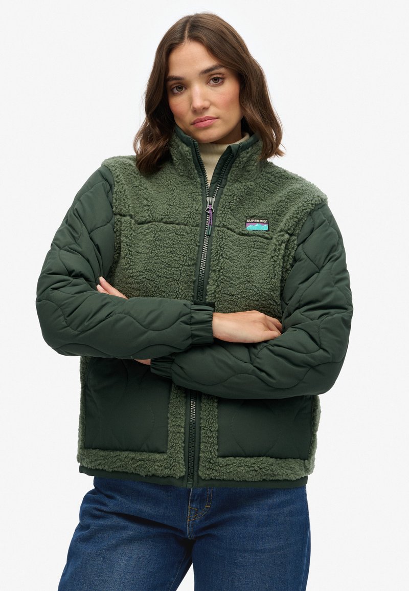 Groene geïsoleerde jas met gequiltte stof en pluizige fleece-textuur. Voorzien van een ritssluiting aan de voorkant, elastische manchetten en een logo-patch op de borst.