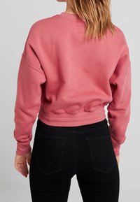 Persona con un sudadera recortada de color rosa polvo y jeans negros de cintura alta, vista desde atrás contra un fondo neutro.