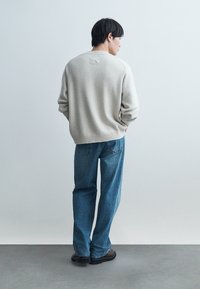 Férfi áll háttal, világosszürke pulóvert, kék farmert és sötét cipőt visel egyszerű világos háttér előtt.