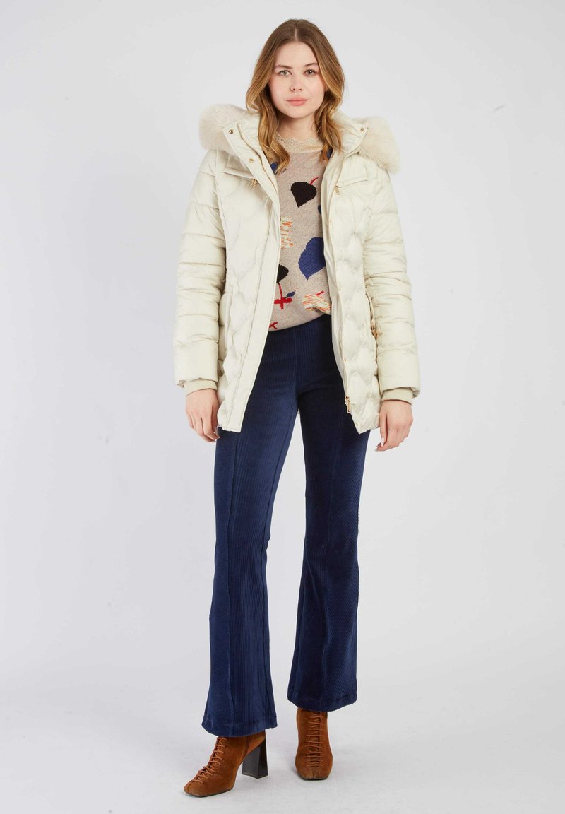 Witte donzen jas met een met bont gevoerde capuchon, gecombineerd met een beige, patroontrui en donkerblauwe corduroy wijde broek. Bruine laarzen met hak.