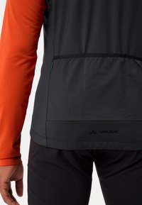 Veste de sport noire et orange avec des manches longues, présentant un design ajusté, un tissu texturé et un logo visible en bas.