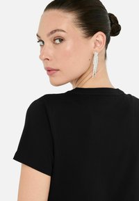 Zwarte t-shirt met korte mouwen, gladde textuur. Opvallende sprankelende druppel oorbellen met langwerpige rhinestone accenten. Profielweergave van een vrouw.