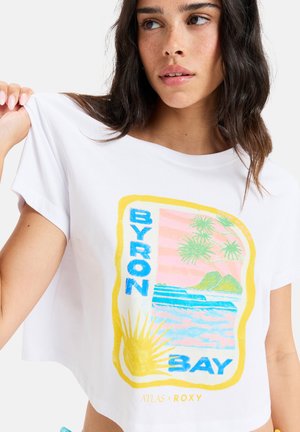 White cropped t-shirt featuring a colorful "Byron Bay" graphic with sun, palm trees, and ocean scene in yellow, pink, blue, and green.