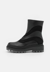 Mustat nilkkurit mid-calf-mittaisilla sekoitettuilla sileillä ja mokkanahkaisilla paneeleilla, paksu chunky-pohja syvillä uriolla ja pyöreä kärki valkoisella taustalla.