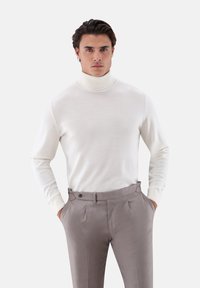 Mann mit dunklen Haaren, der einen weißen Rollkragenpullover und beige hoch taillierte Anzughosen trägt und die Hände in den Taschen hat.