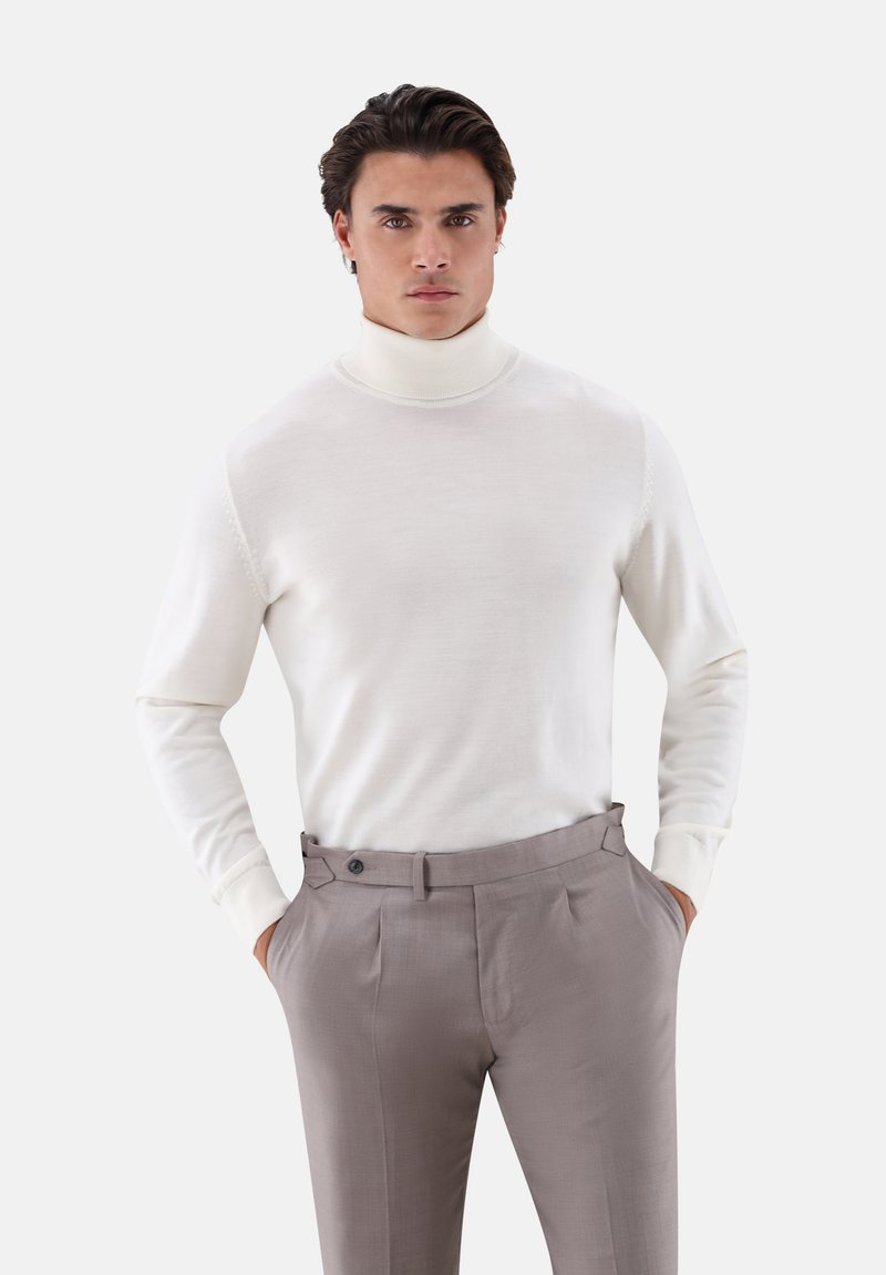Mann mit dunklen Haaren, der einen weißen Rollkragenpullover und beige hoch taillierte Anzughosen trägt und die Hände in den Taschen hat.
