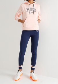 Kvinna som bär en ljusrosa Nike-hoodie, mörkblå Nike leggings med ränder, och vita sneakers med orange sulor, stående med händerna i fickorna.