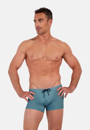 Muskelöser Mann mit kurzen braunen Haaren, der blaue gemusterte Badehosen mit schwarzer Kordel trägt, steht mit den Händen in den Hüften und schaut nach links.