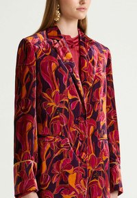 Veste en velours à motif floral dans des tons de violet, rose et orange. Comprend des revers crantés, deux poches avant et une fermeture par boutons.