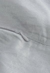 Šedá tkanina s jemnou texturou a leskem. Na horním okraji se nachází malý zip v světlé šedé barvě, začleněný do materiálu.