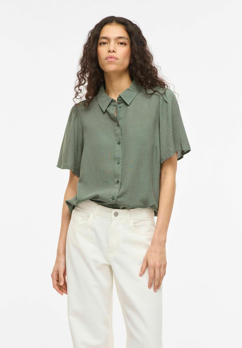 Jeune femme aux cheveux bouclés portant une chemise verte ample à boutons et un pantalon blanc taille haute, debout devant un fond uni.