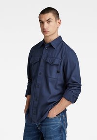 Chemise à manches longues bleu marine, dotée de deux poches poitrine, d'une fermeture à boutons et de manches retroussées. Fabriquée en tissu lisse sans motifs visibles.