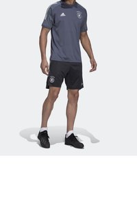 Szara sportowa koszulka z czarnymi spodenkami, z trzema białymi paskami, logo oraz okrągłą naszywką z gwiazdkami. Czarne buty sportowe dopełniają wygląd.