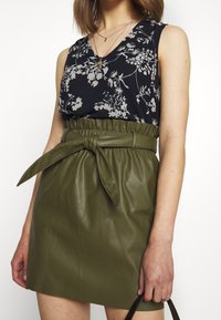 Zwart bloemenpatroon zonder mouwen top, gecombineerd met een hoge taille, olijfgroen nepleer rok met een strikceintuur detail.