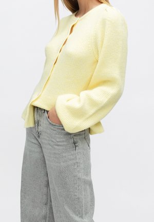 Femme portant un cardigan tricoté jaune clair avec des manches bouffantes, associé à un jean taille haute gris clair, main dans la poche, sur un fond uni.