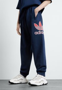 Calças de moletão azul-marinho com cintura elástica e bolsos laterais, apresentando um logo da Adidas em vermelho e branco na coxa esquerda. Ténis brancos usados.