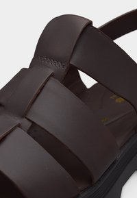 Καφέ δερμάτινο σανδάλι με φαρδιές πλεγμένες λουρίδες και εμφανές ράψιμο σε μαύρη σόλα, λεπτομέρεια κοντινής λήψης.