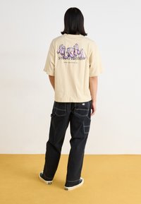 T-shirt en coton beige avec un imprimé graphique dans le dos ; associé à un pantalon cargo noir avec des coutures blanches et des poches.