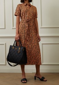 Robe à imprimé léopard orange avec manches courtes et ceinture à la taille, accompagnée d'un sac à main noir matelassé avec des accents dorés et des sandales noires à brides.