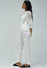 Top de lino blanco con detalles de encaje en las mangas, combinado con pantalones de lino blanco y sandalias negras con acentos dorados.