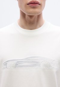 Λευκή φαρδιά μπλούζα με διακριτική, κεντημένη σιλουέτα αυτοκινήτου στο στήθος, που παρουσιάζει ομαλές γραμμές και μινιμαλιστικό σχεδιασμό.