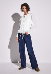 Camicia bianca a maniche lunghe con tessuto con texture e vita regolabile, abbinata a jeans in denim blu scuro a gamba dritta e scarpe marroni.
