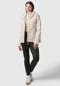 Lichtbeige waterdichte jas met een capuchon, knoopaccenten en een koord in de taille, gedragen over een gestreept shirt en zwarte slim-fit broek.