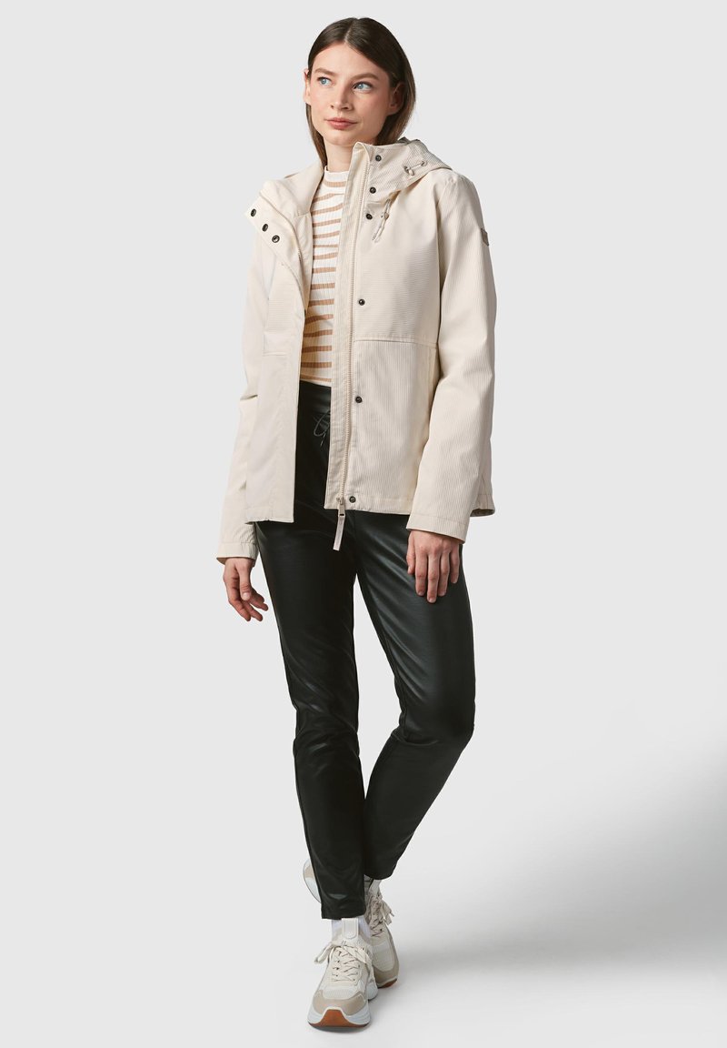 Lichtbeige waterdichte jas met een capuchon, knoopaccenten en een koord in de taille, gedragen over een gestreept shirt en zwarte slim-fit broek.