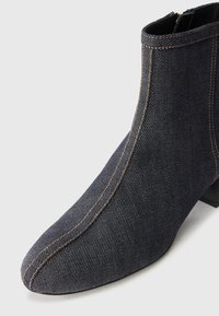 Bota de tornozelo em denim escuro com bico apontado, costura amarela contrastante e um salto baixo em bloco. Textura suave e design elegante.