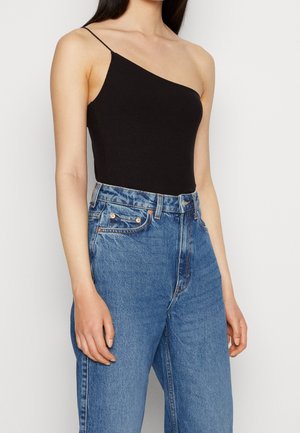 Femme portant un haut noir côtelé à une épaule avec une fine bretelle et un jean bleu taille haute sur un fond uni.