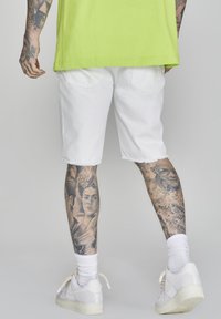 Shorts di denim bianco con orli sfrangiati, abbinati a una maglietta over-size verde brillante e scarpe da ginnastica bianche. Tatuaggi visibili sulle gambe.