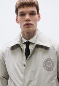 Ανοιχτό γκρι μπουφάν με καλοσχηματισμένο γιακά, κλείσιμο με φερμουάρ και διακοσμητική λεπτομέρεια. Φορεμένο πάνω από λευκό πουκάμισο και μαύρη γραβάτα. Λείο υλικό.