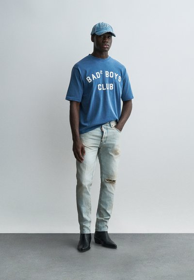 Homme portant un t-shirt bleu "BAD² BOYS CLUB", un jean délavé clair avec déchirure au genou, des bottes noires et une casquette bleue, debout contre un mur clair uni.