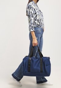 Tmavě modrá texturovaná sportovní taška s černými prvky, vybavená dvojitými úchyty a venkovními kapsami, kterou nese osoba v neform álním oblečení.