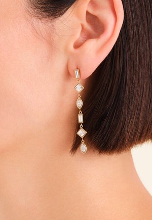 Boucle d'oreille en or pendante avec des pierres précieuses transparentes de forme rectangulaire, carrée et marquise sur une personne aux cheveux courts et foncés.