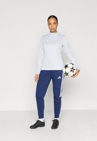 Ανοιχτό μπλε αθλητικό μακρυμάνικο μπλουζάκι με φερμουάρ στο γιακά. Σκούρο μπλε παντελόνι φόρμας με λευκές ρίγες και λογότυπο Adidas. Μαύρη ποδοσφαιρική μπάλα που κρατιέται στο χέρι.