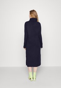 Robe en maille bleu marine à col roulé avec manches longues, fentes latérales et poignets côtelés, associée à des bottines à talons vert vif.