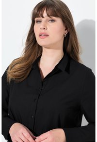 Černá dlouh�á košile s límečkem na knoflíky, předními knoflíky a hladkým povrchem. Model pózuje s rukama u pasu na světlém pozadí.