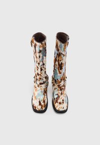Botas de caña alta marrón y blanca con un acabado brillante, que cuentan con dos hebillas y una suela negra. Textura suave con un acento brillante.